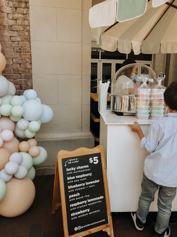 Cotton candy umbrella cart set up next to pastel balloon arch with chalkboard menu in front | Whim Cotton Candy cart at Shop Hop on Palafox Street