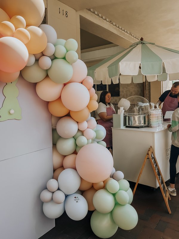 Cotton candy umbrella cart set up next to pastel balloon arch with chalkboard menu in front | Whim Cotton Candy cart at Shop Hop on Palafox Street