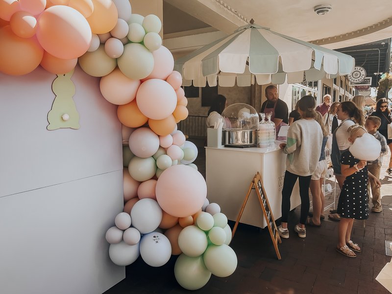 Cotton candy umbrella cart set up next to pastel balloon arch with chalkboard menu in front | Whim Cotton Candy cart at Shop Hop on Palafox Street