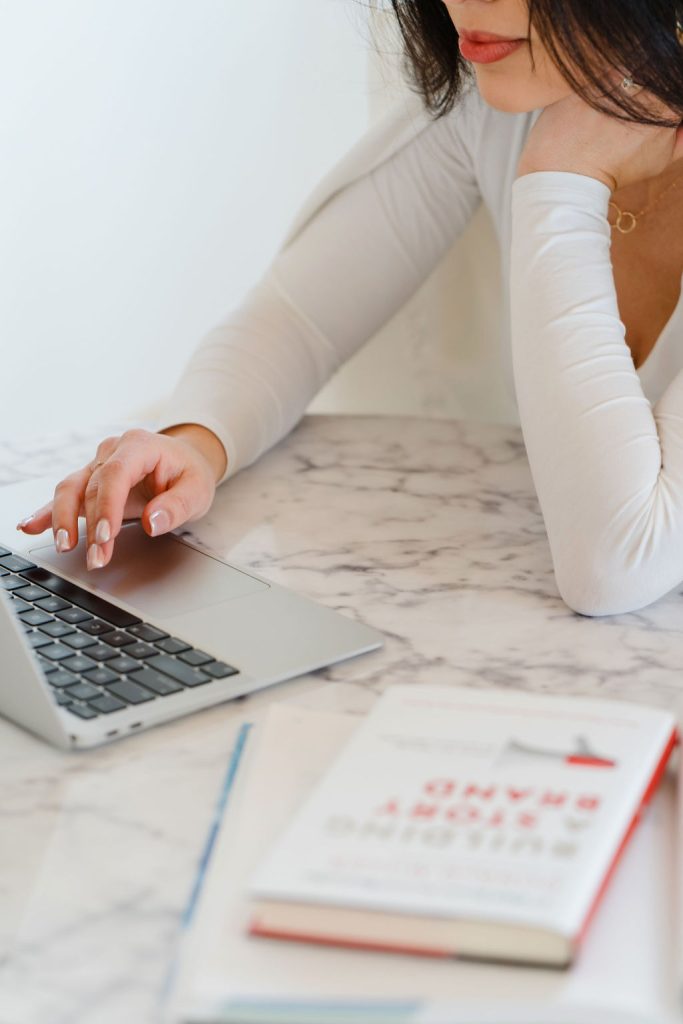 Woman sits at a round marble cafe table and types on a MacBook air beside a copy of the book 
How to Build A StoryBrand"