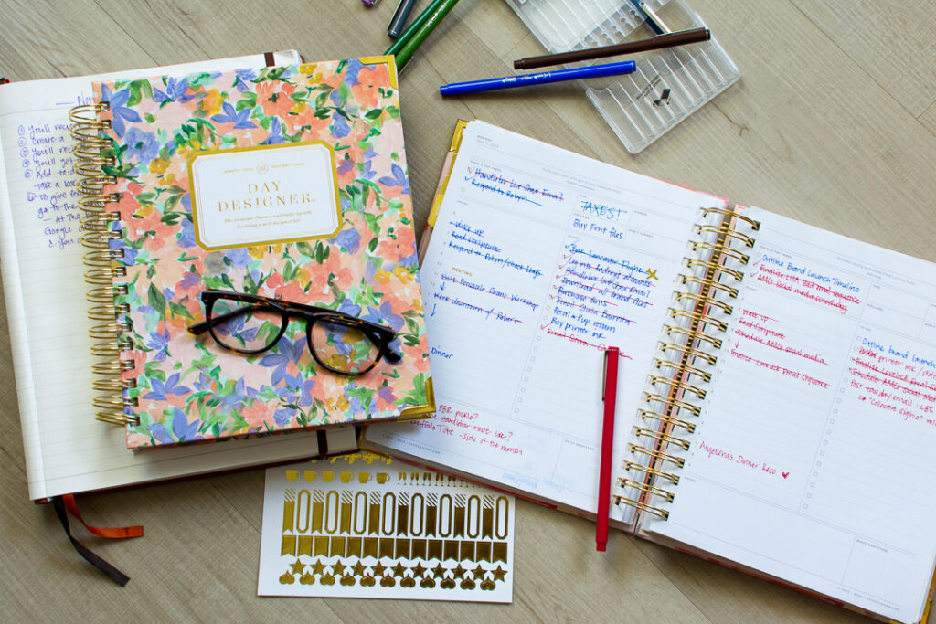 Floral print Day Designer planner sits next to open page of daily planner next to multi colored pens and a gold sticker sheet | Lifestyled By Sofia