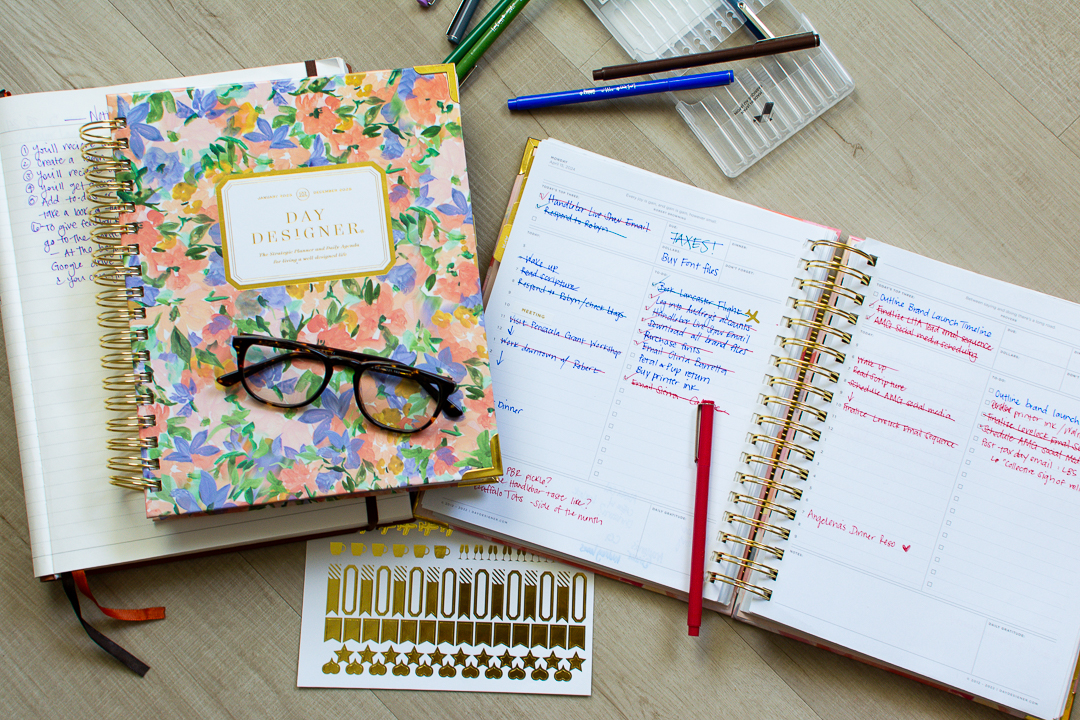 Floral print Day Designer planner sits next to open page of daily planner next to multi colored pens and a gold sticker sheet | Lifestyled By Sofia