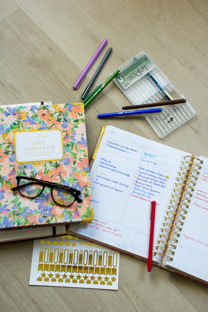 Floral print Day Designer planner sits next to open page of daily planner next to multi colored pens and a gold sticker sheet | Lifestyled By Sofia
