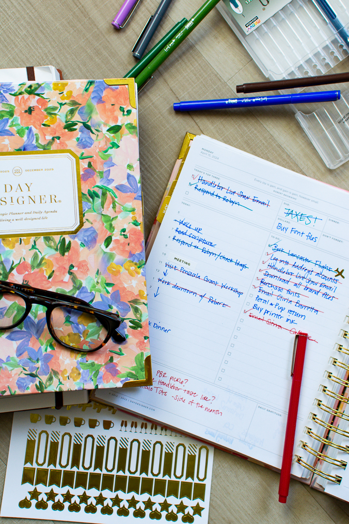 Floral print Day Designer planner sits next to open page of daily planner next to multi colored pens and a gold sticker sheet | Lifestyled By Sofia