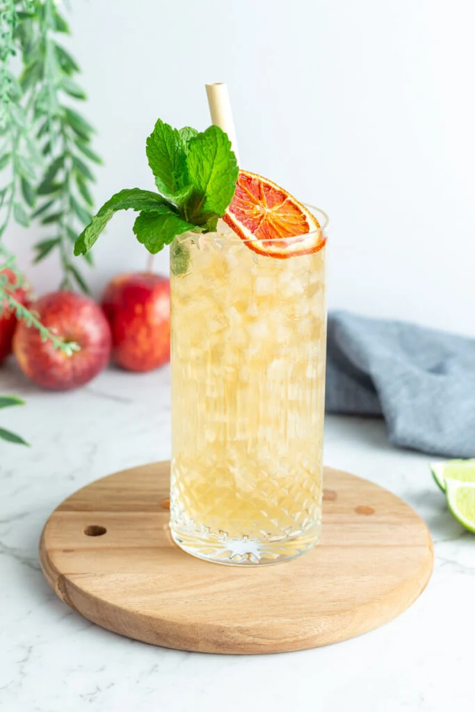 Apple Ginger Fizz Mocktail in a tall glass sits on a round wooden tray with apples behind it | Seasonal Mocktails roundup by Lifestyled By Sofia