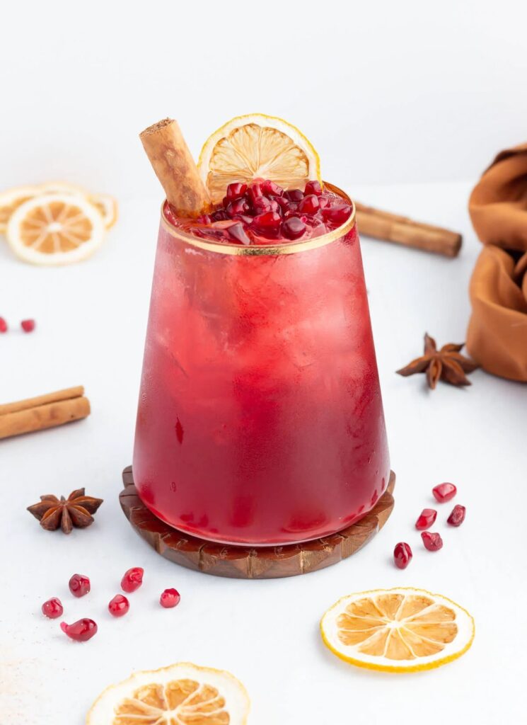 Pomegranate Cinnamon Mocktail in a gold rimmed glass surrounded by pomegranate seeds and cinnamon sticks | Festive Thanksgiving Mocktails roundup by Lifestyled By Sofia