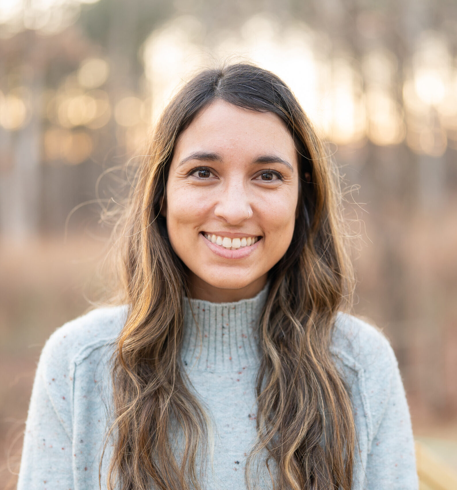Woman with long brown wavy hair smiles with sun setting in the trees in the background