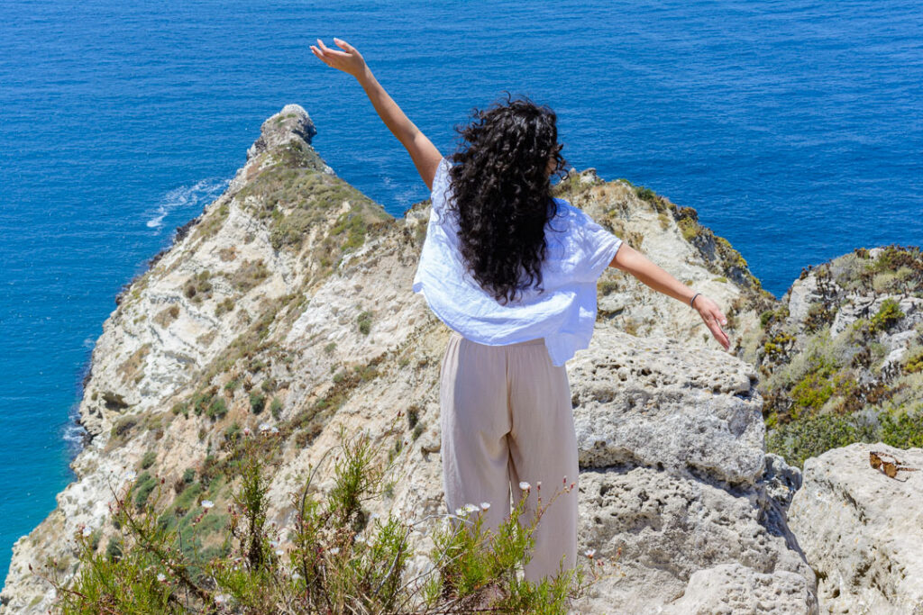 Woman in linen pants and shirt stands with arms outstretched on a rocky cliff overlooking blue waters on Santa Cruz Island, Channel Islands National Park