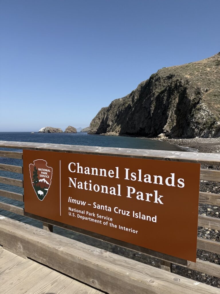 Channel Islands National Park entrance sign for Santa Cruz Island on the pier, with sea cliffs and a cove in the background