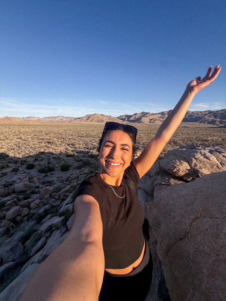 Smiling woman taking a selfie with one arm raised overlooking the desert valley at sunset on the Bigfoot Panorama trail in Joshua Tree National Park